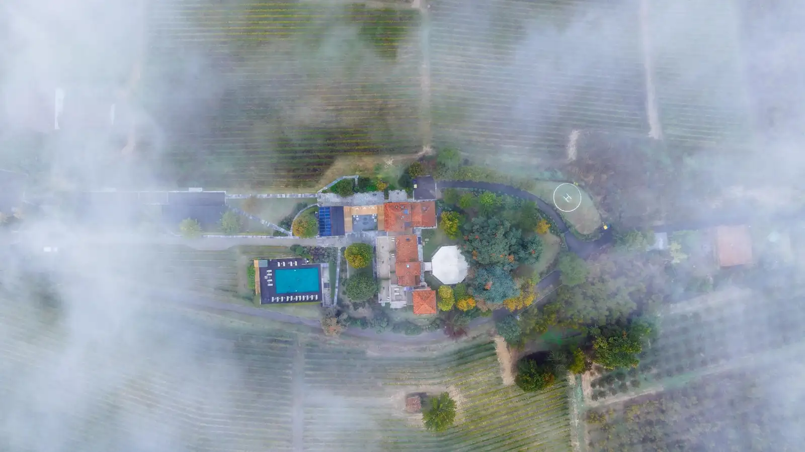 Drone shot capturing a villa with swimming pool amidst foggy fields in Piemonte, Italy.
