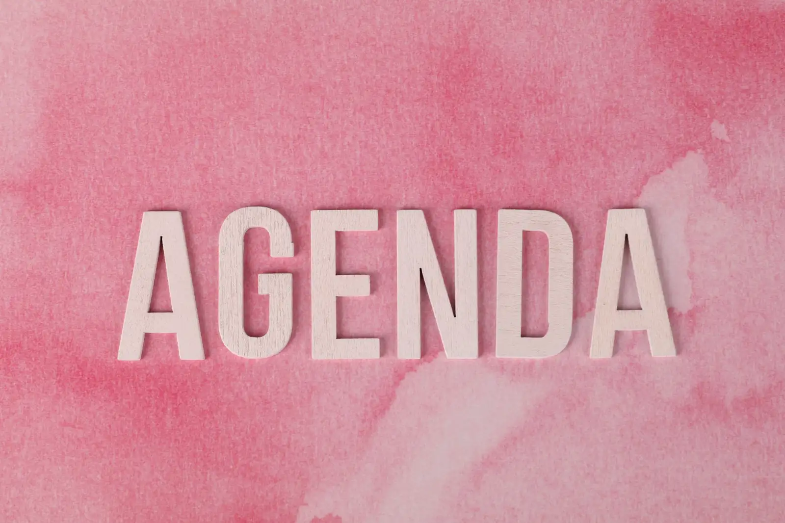 Wooden letters forming the word 'AGENDA' on a pink watercolor background.