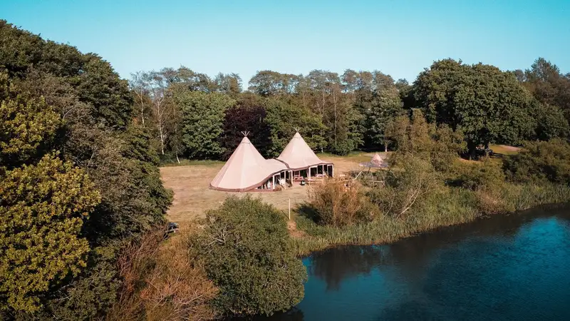 A serene aerial view of tipis beside a lake surrounded by lush forest during summer.